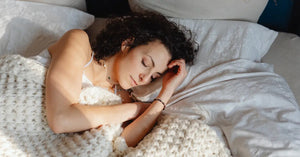 CBD for sleep helps a woman with curly hair rest peacefully wrapped in white bedding and a knit throw.