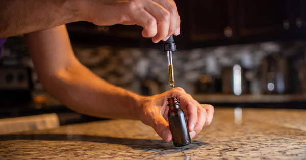 CBD tincture being dispensed from a glass dropper into an amber bottle on a granite kitchen countertop.