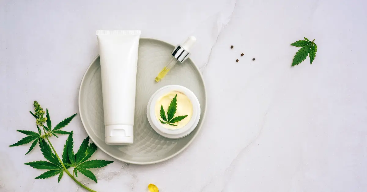 Best topical CBD products displayed with a cream jar, dropper, and tube on a plate with hemp leaves.